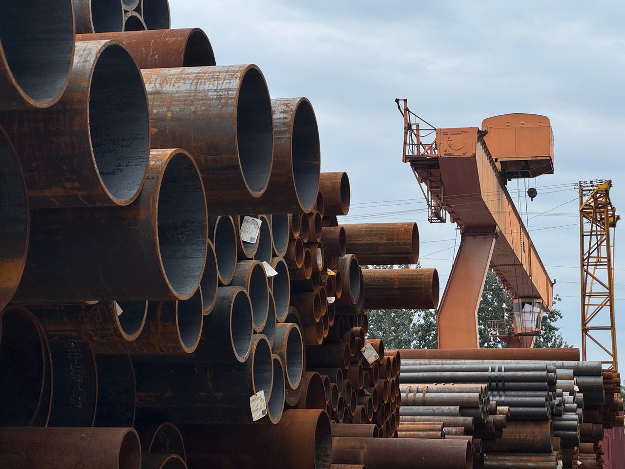 services-03 Stacked steel pipes beside an industrial crane in an outdoor yard, showcasing heavy industry and construction materials.